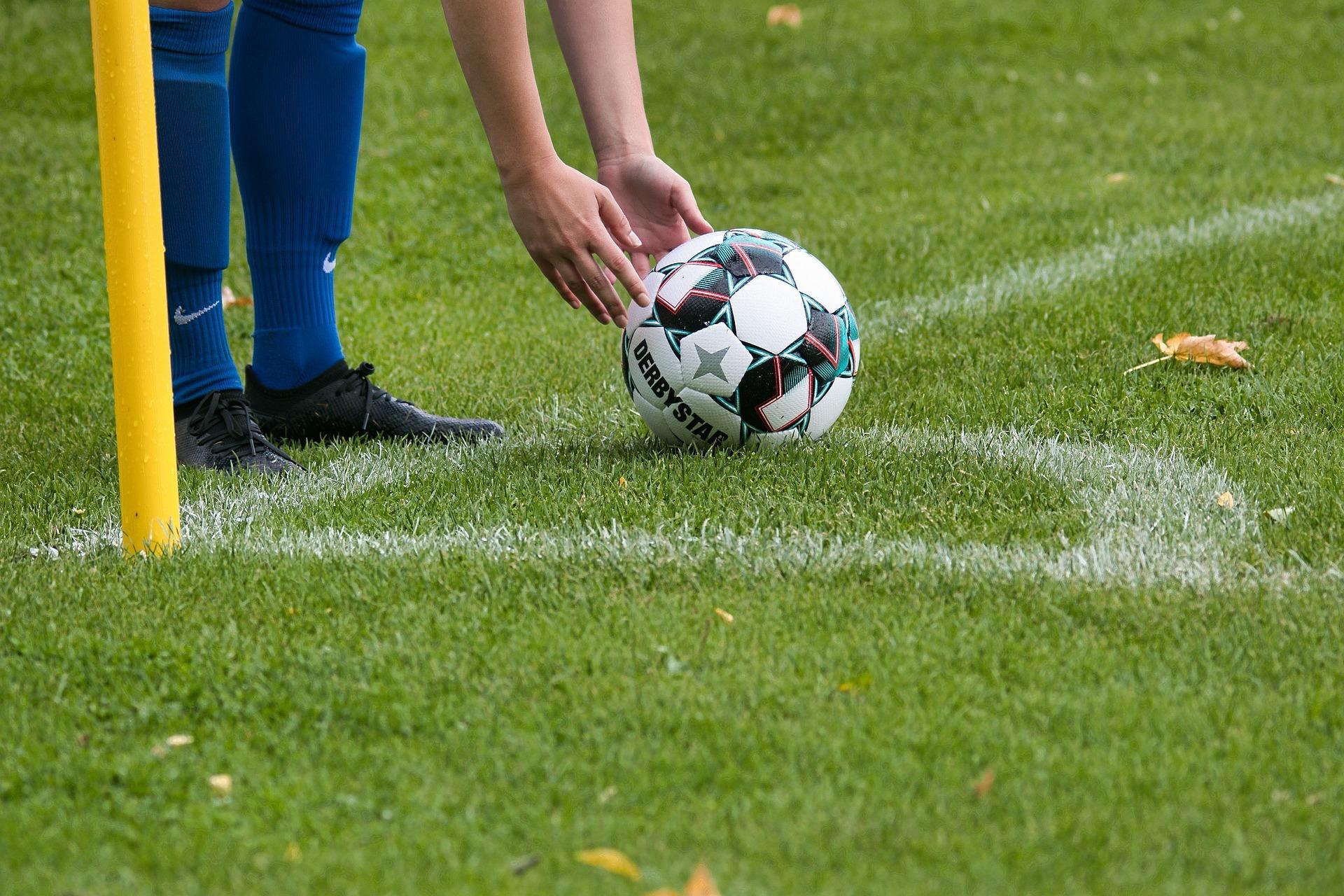 Hoek van een voetbalveld. In beeld zwarte voetbalschoenen en blauwe sokken en handen die een bal neerleggen.