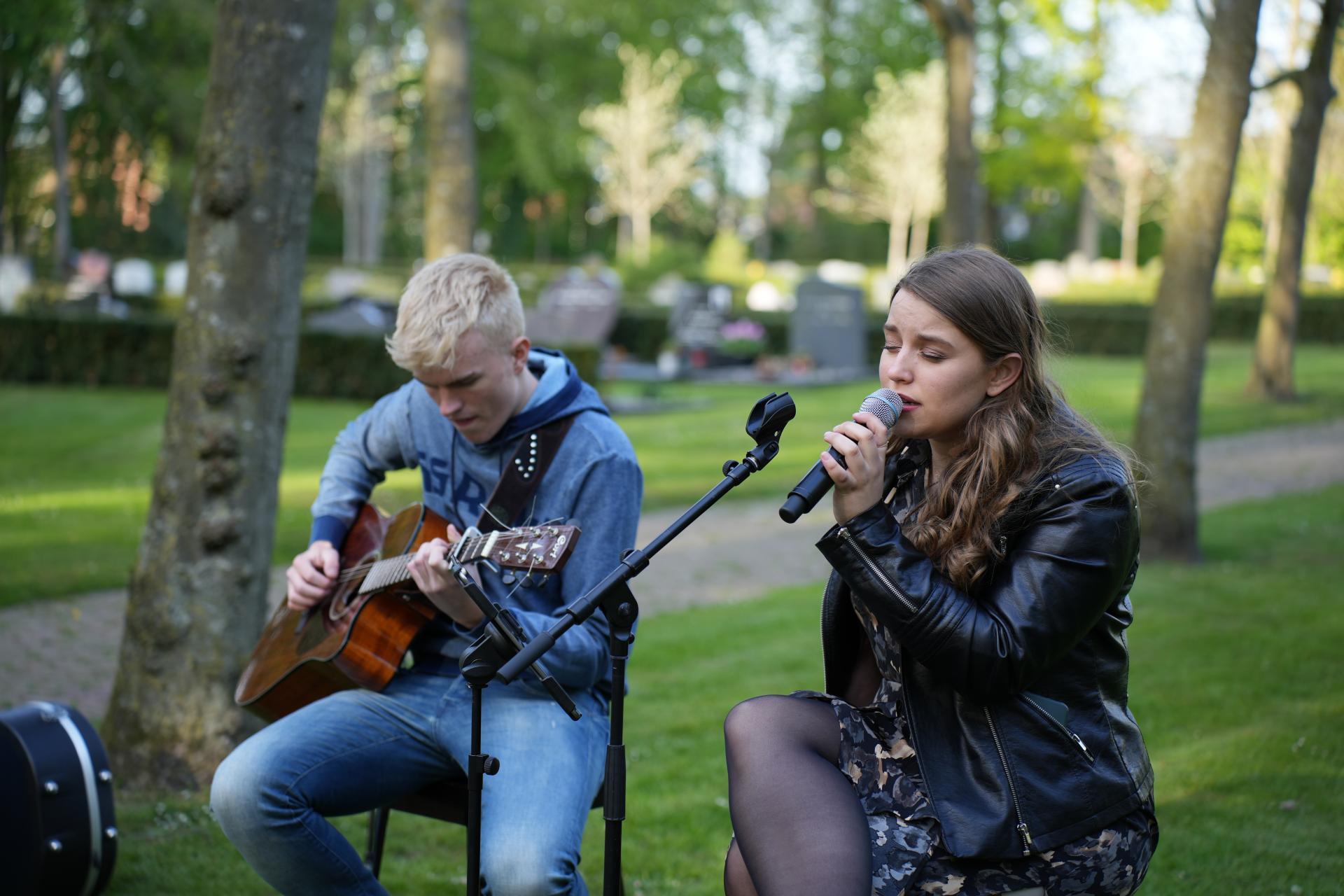 Een jongen die op een gitaar speelt en een meisje dat in een microfoon zingt. Ze zitten op een stoel op een begraafplaats. Ver op de achtergrond graven.