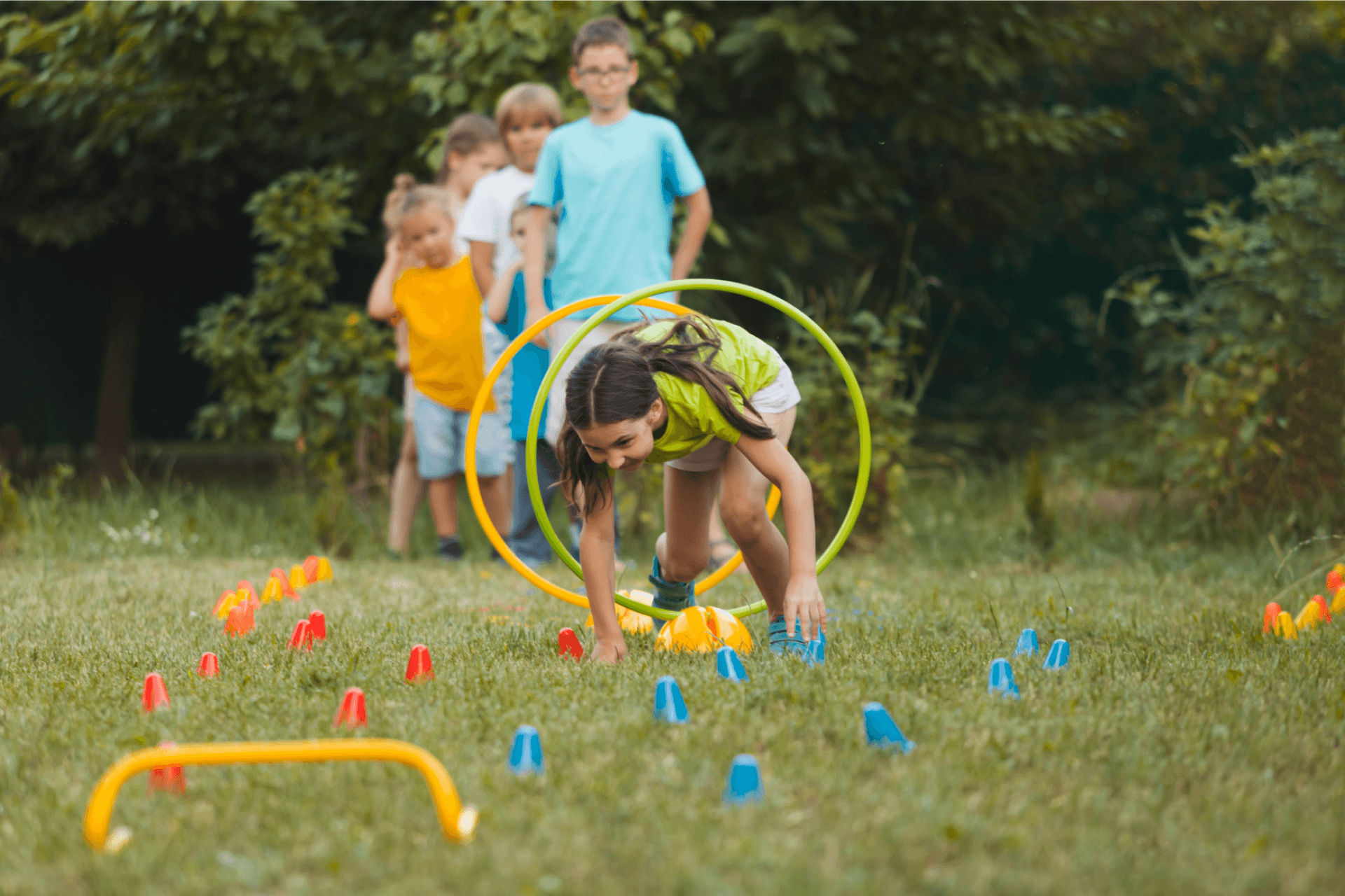 Een kind kruipt door een rechtopstaande hoepel op een grasveld. Achter het kind mee hoepels en voor haar gekleurde blokjes. Achter de hoepels staat een rijtje kinderen.